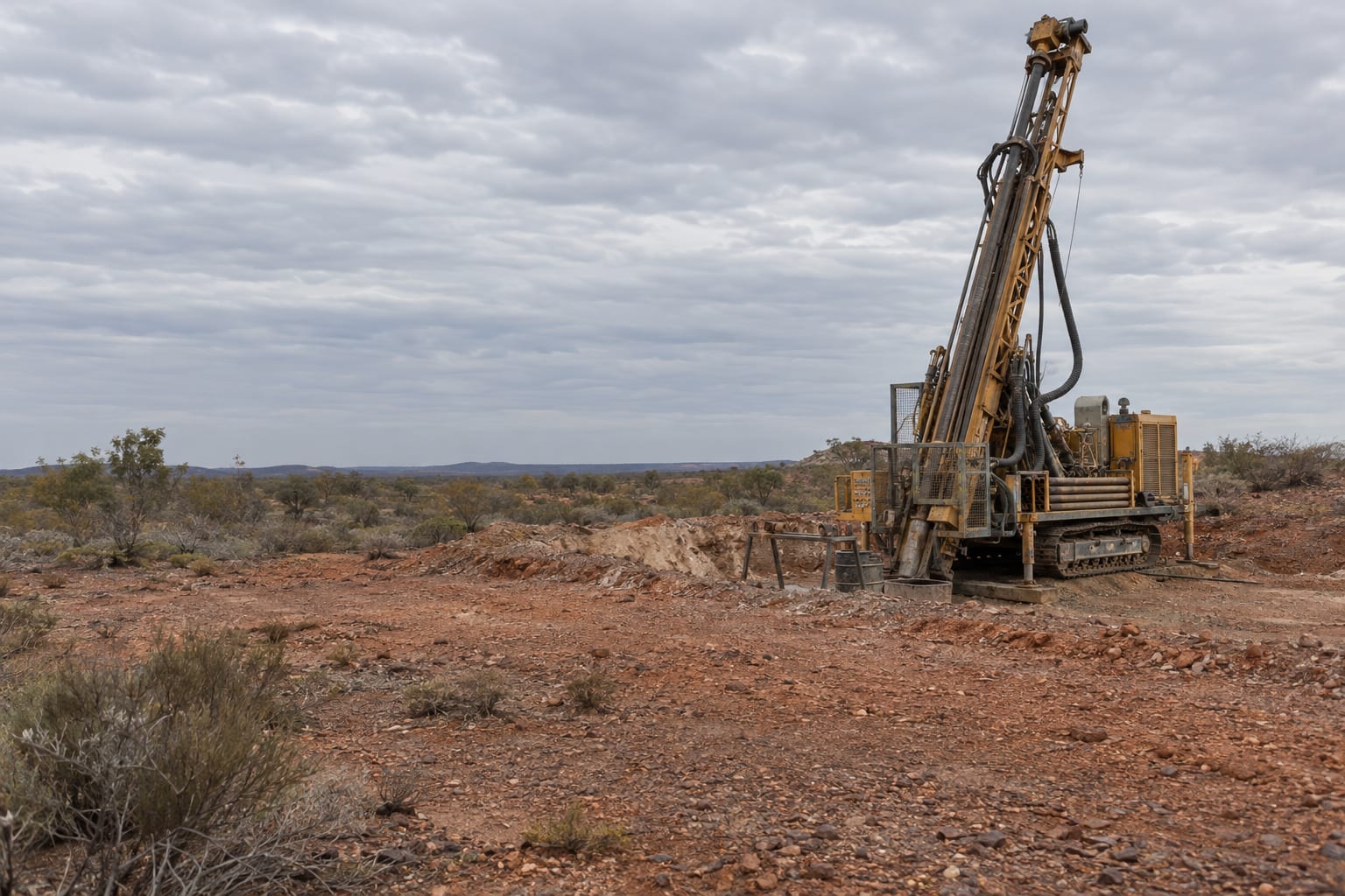 A parked, unbranded drill rig stands on red gravel beneath an overcast sky at an anonymous goldfield lease.
