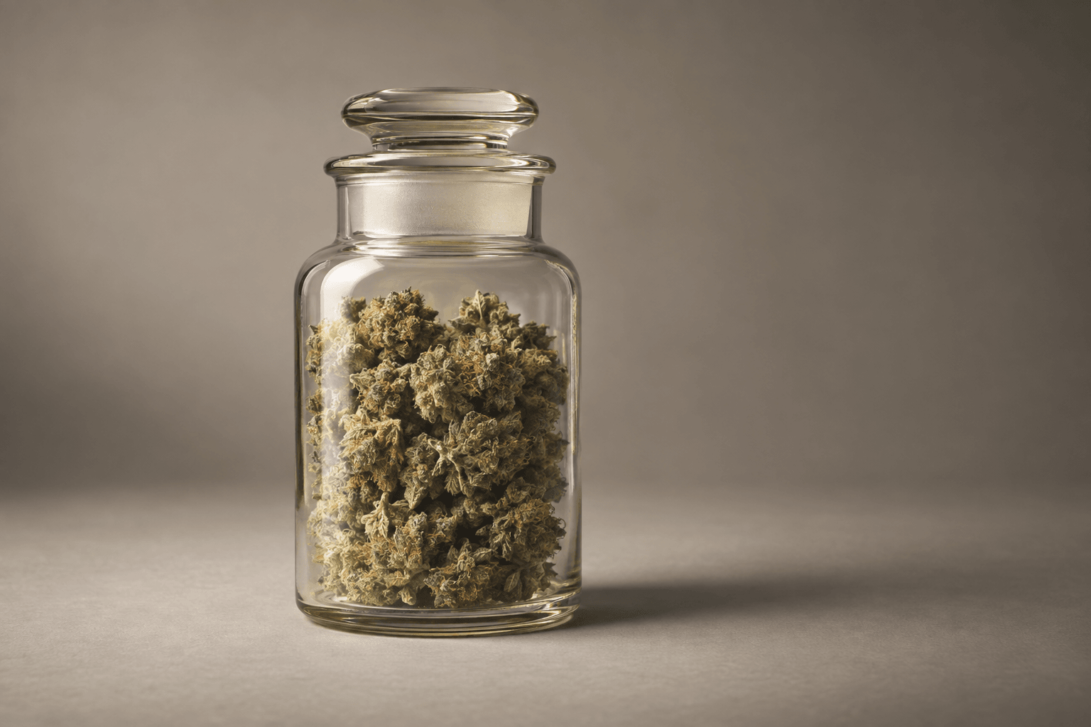 A sealed glass jar of dried cannabis flower stands on a pale seamless backdrop under studio light.