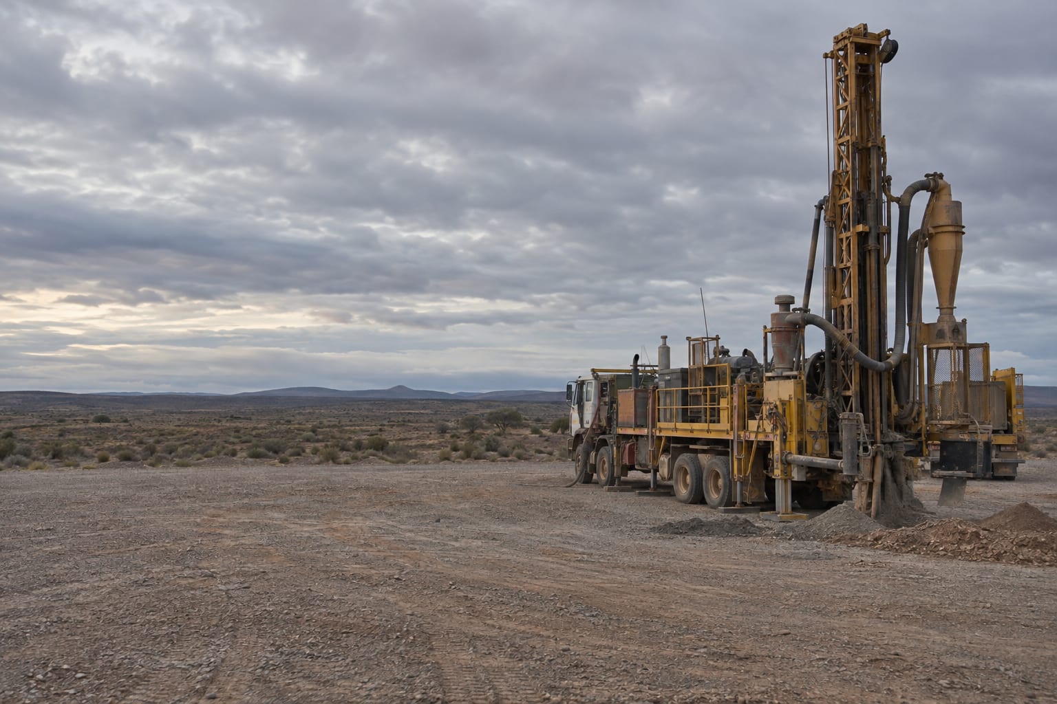 A reverse circulation drill rig stands on a gravel exploration pad under an overcast sky.