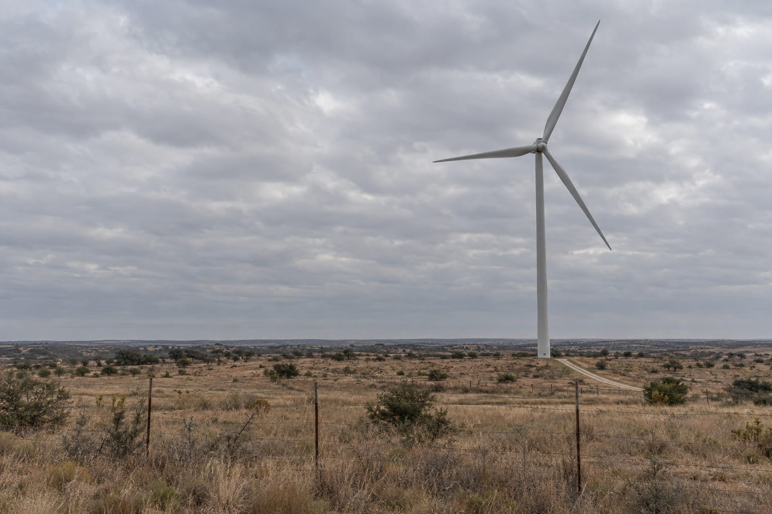 A wind turbine rises above dry grassland beneath an overcast sky.