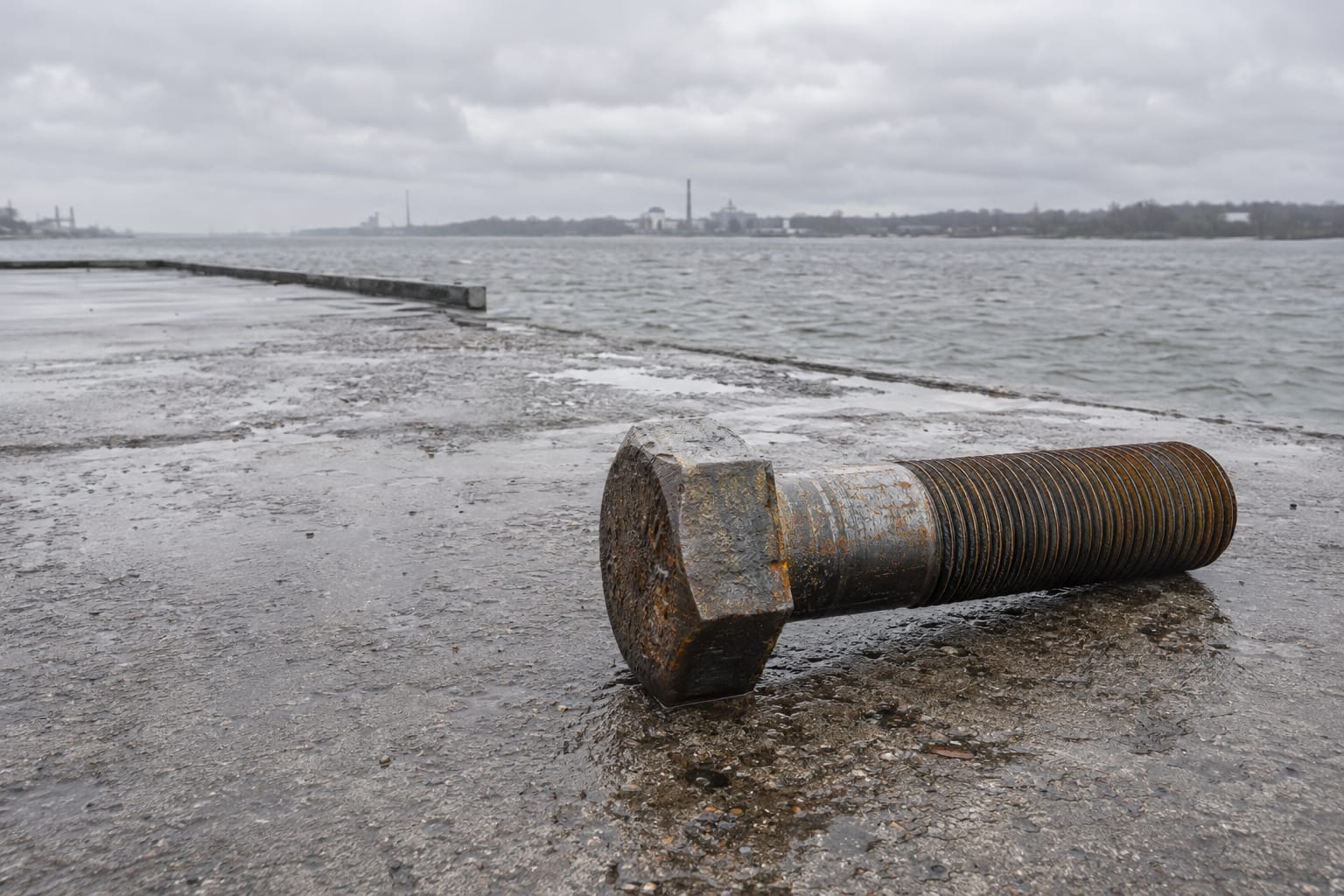 An oversized turbine bolt rests on an anonymous freight quay beside a grey river.