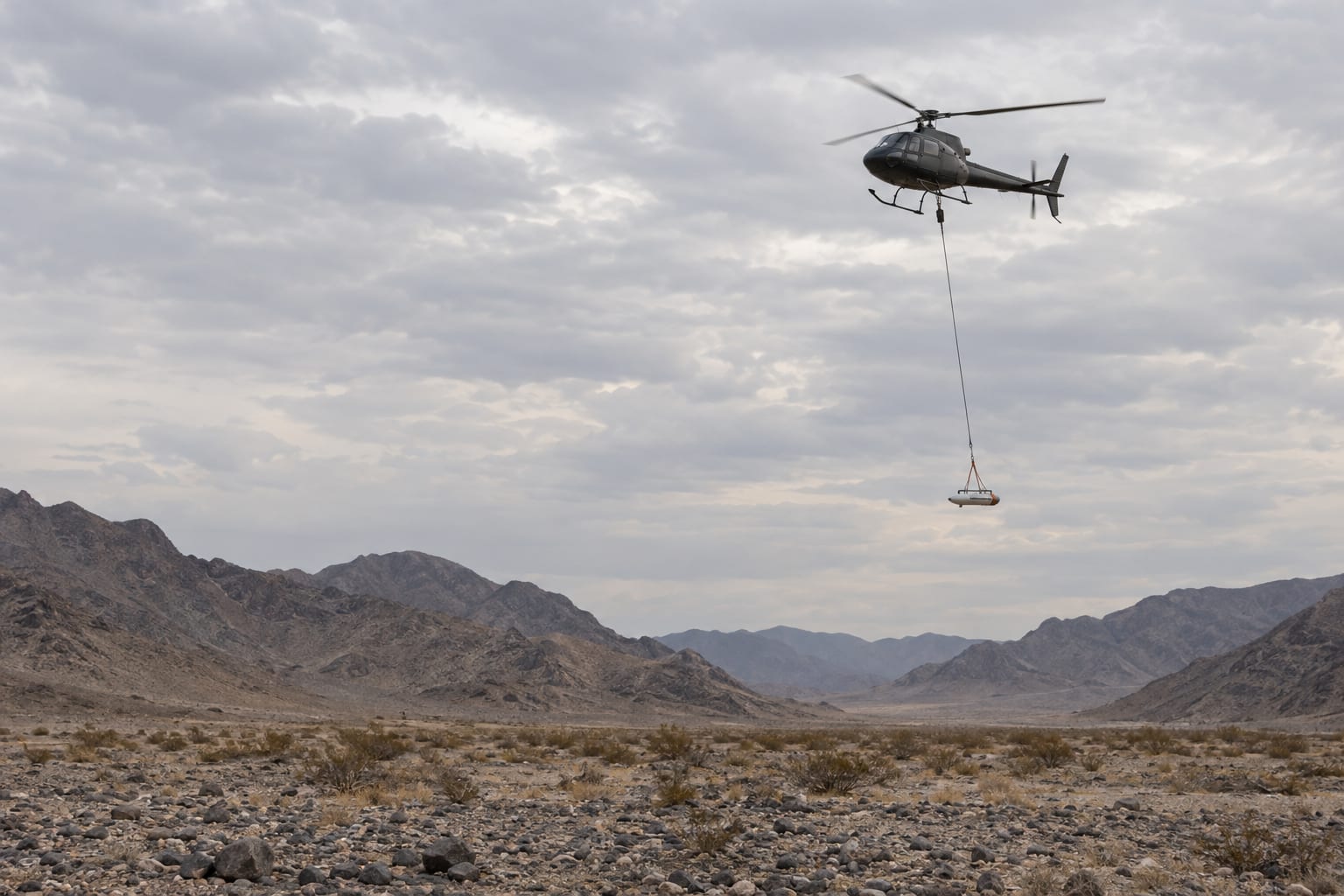 An unmarked helicopter tows an airborne geophysical sensor above a generic desert basin under overcast skies.