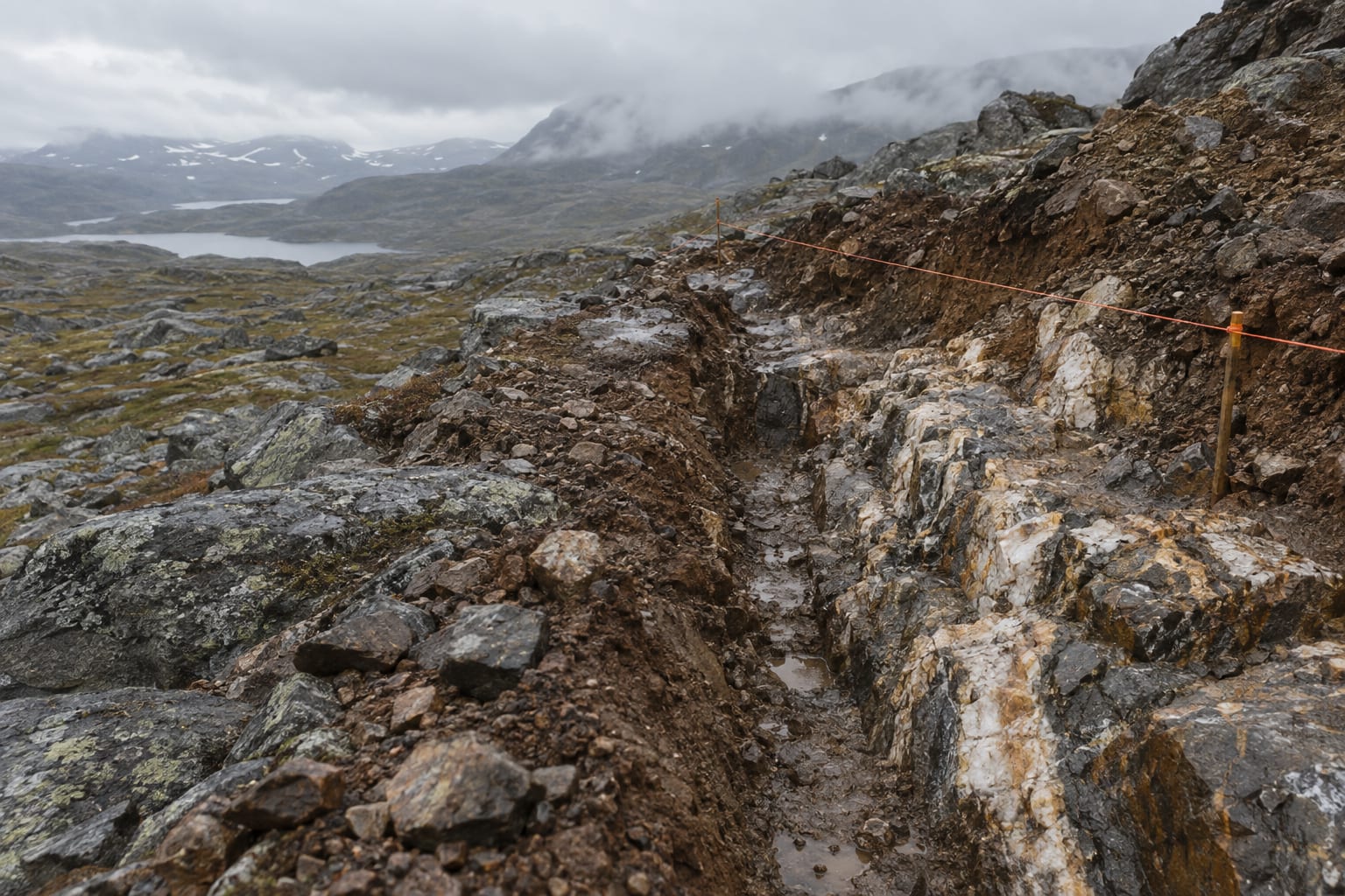 A rain-darkened exploration trench exposes quartz-veined bedrock on a rocky upland.
