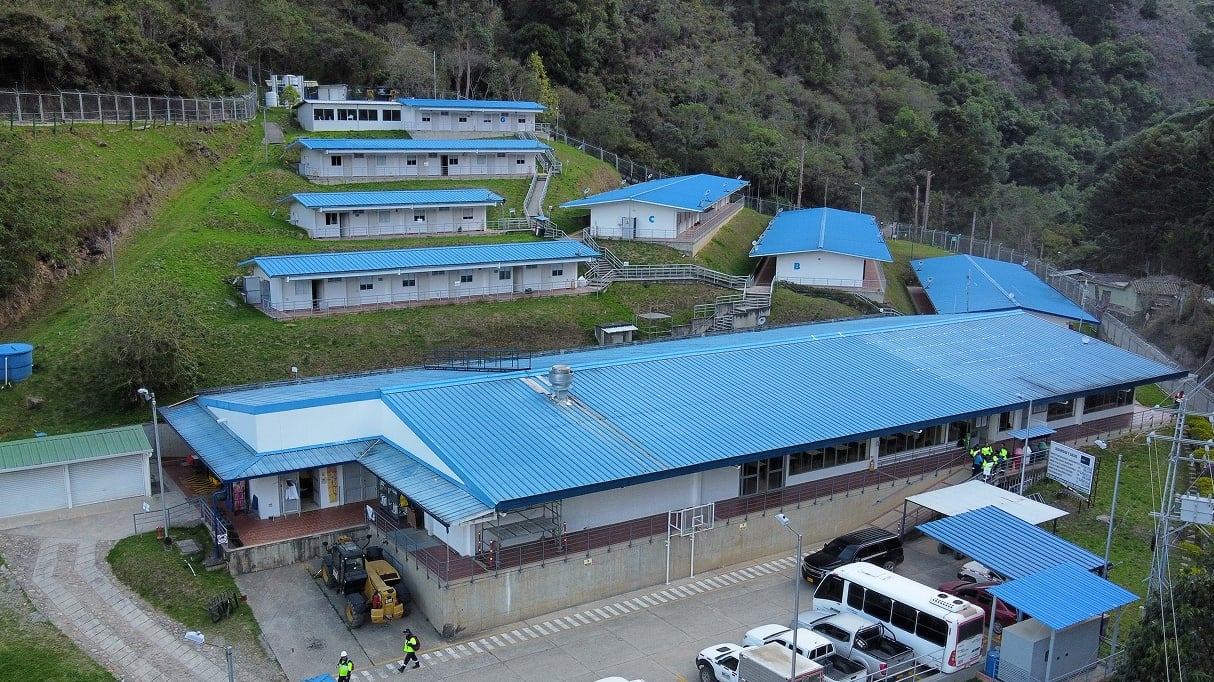 Blue-roofed industrial buildings stand among green hills, with vehicles and workers visible in front.