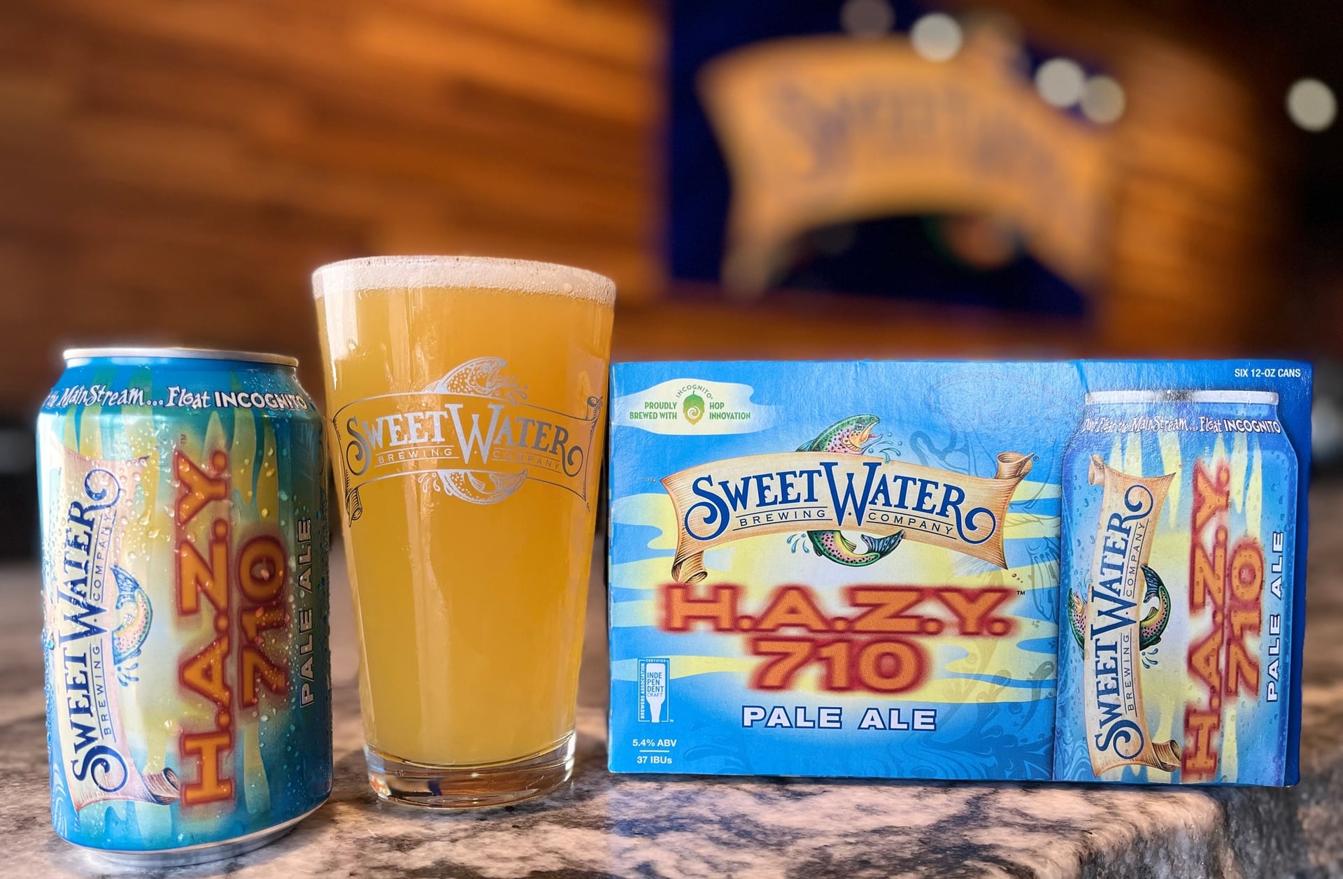 A single SweetWater's Hazy 710 Pale Ale can next to a glass of beer and case of the Pale Ale